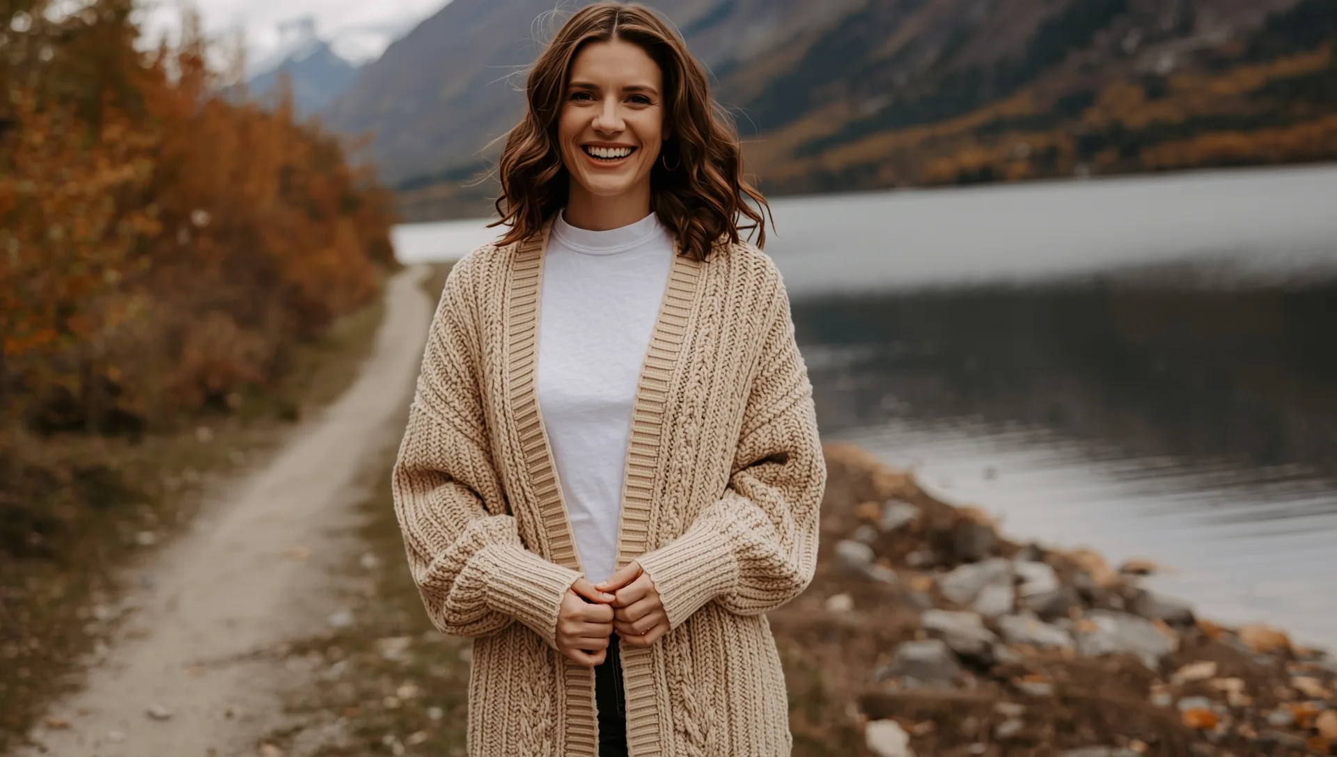 Smiling woman wearing an oversized chunky open-front cardigan on a mountain lake trail